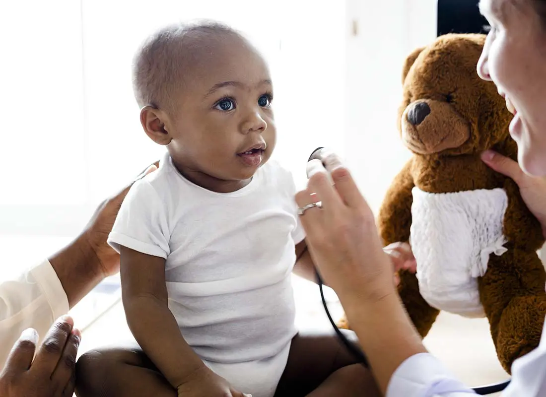 Individual-Health-Insurance-Baby-during-a-Doctor-Visit-for-a-Wellness-Checkup-with-the-Doctor-Holding-a-Childs-Teddy-Bear-to-Try-and-Keep-Childs-Attention-and-to-Stay-Positive