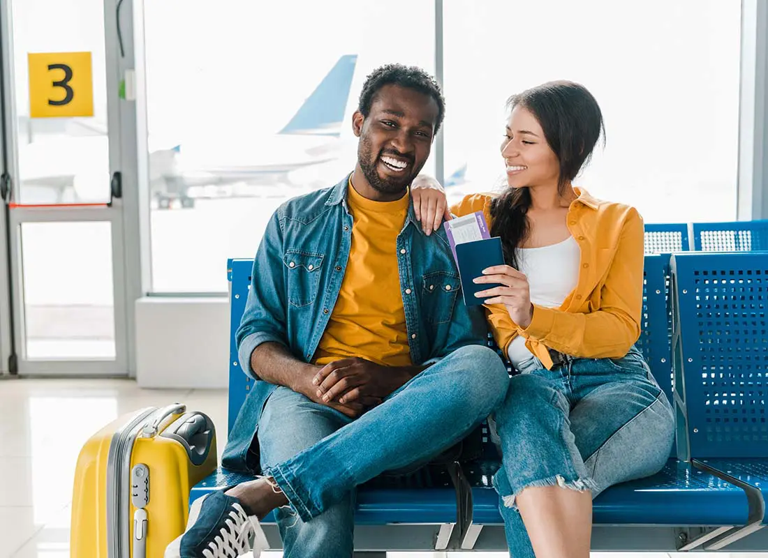 Travel-Insurance-Smiling-Young-Couple-Sitting-in-the-Departure-Lounge-with-Baggage-and-Tickets-in-the-Airport-with-Airplane-behind-in-the-Distance